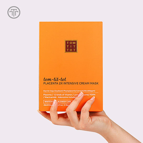 Orange box of tom-tit-tot Placenta 2X Intensive Cream Mask held by a hand on a light gray background